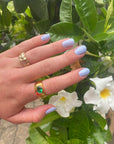 Close-up of a hand with light lavender nails painted with M+M - Clean Nail Polish, adorned with gold rings, gently resting near white flowers and green leaves outdoors.