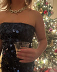 Woman wearing a black sequin dress and gold chain necklace holding a glass, showcasing nails painted with Gold Foil - Clean Nail Polish in front of a decorated Christmas tree.