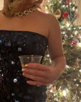 Woman with metallic gold nails wearing a black sequined dress holding a clear glass in front of a decorated Christmas tree showcasing Gold Foil - Clean Nail Polish.