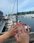 Hands with polished coral-red nails using Candlewood Coral - Clean Nail Polish, adorned with rings and a bracelet, posed over a marina with boats and calm water.